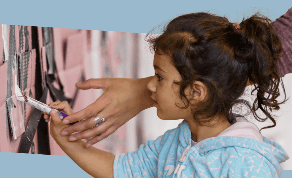 A young girl paints a large wall mural with an adult’s guiding hand, concentrating intently in a collaborative art activity.